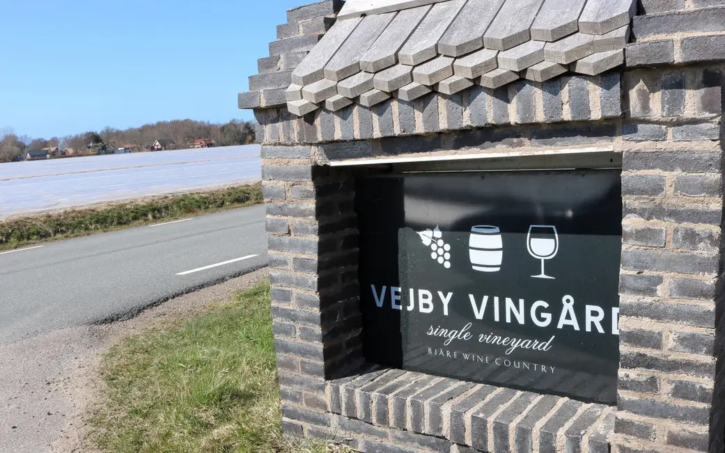 En tegelbyggd vägskylt vid infarten till Vejby Vingård, med texten 'single vineyard' och symboler för druvor, ett vinfat och ett vinglas. I bakgrunden syns öppna fält och en landsväg under en klarblå himmel.